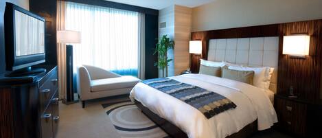 A hotel room with a large bed, a desk with a flat-screen TV, a chair, a plant, and a nightstand with a lamp.