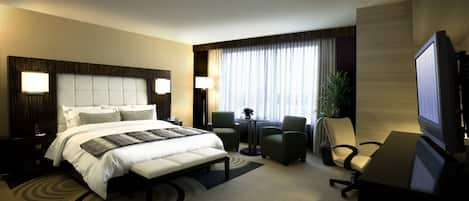A hotel room with a large bed, two armchairs, a desk with a computer, and a TV.