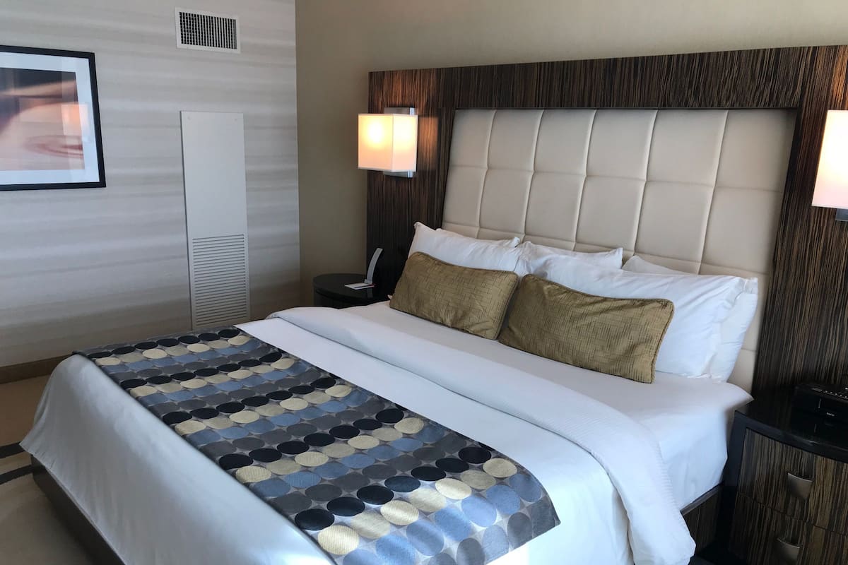 A hotel room with a large bed, a bedside table, a lamp, and a wall-mounted air vent.