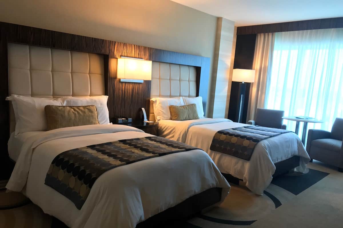 A hotel room with two beds, a nightstand, a lamp, a chair, and a view of the outdoors.