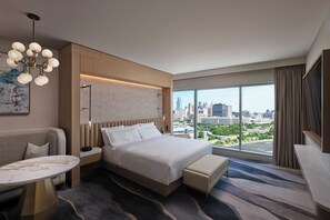 Una habitación de hotel moderna con una cama grande, una zona de estar con una mesa y vistas a la ciudad a través de amplios ventanales.