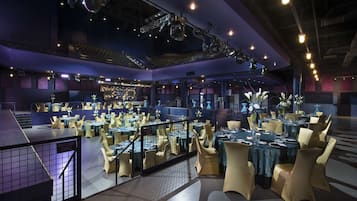 Un amplio comedor interior con varias mesas y sillas, un escenario con equipo de iluminación, y un balcón con barandilla.