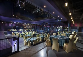 Un amplio comedor interior con varias mesas y sillas, un escenario con equipo de iluminación, y un balcón con barandilla.