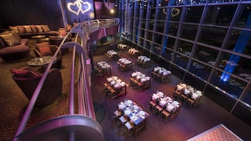 A spacious, multi-level restaurant with a modern design, featuring a variety of seating options including tables and sofas, and large windows offering views of the city.