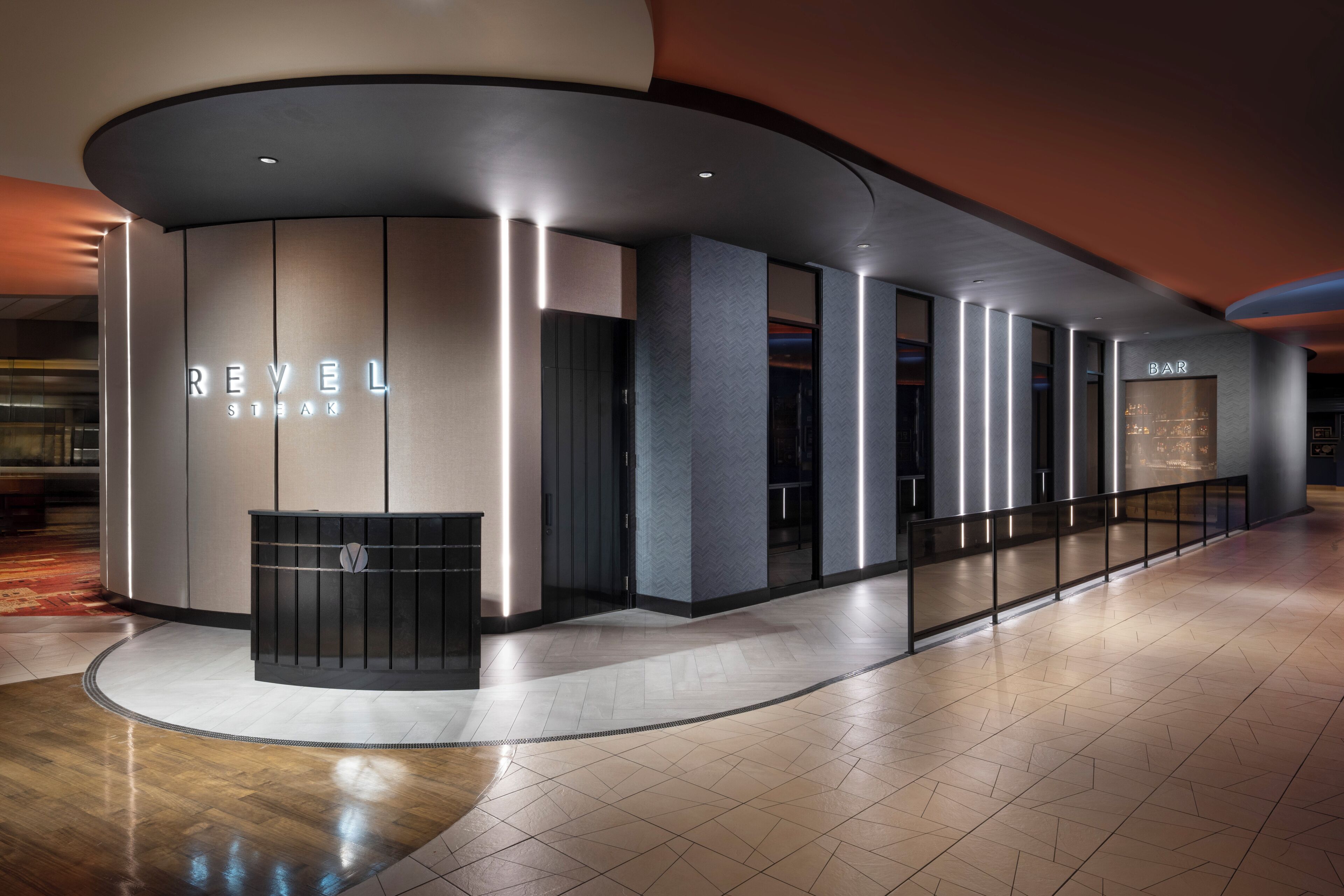 Un hall d’hôtel moderne avec un comptoir d’accueil incurvé portant l’inscription « REVEL STEAK » et un espace bar.
