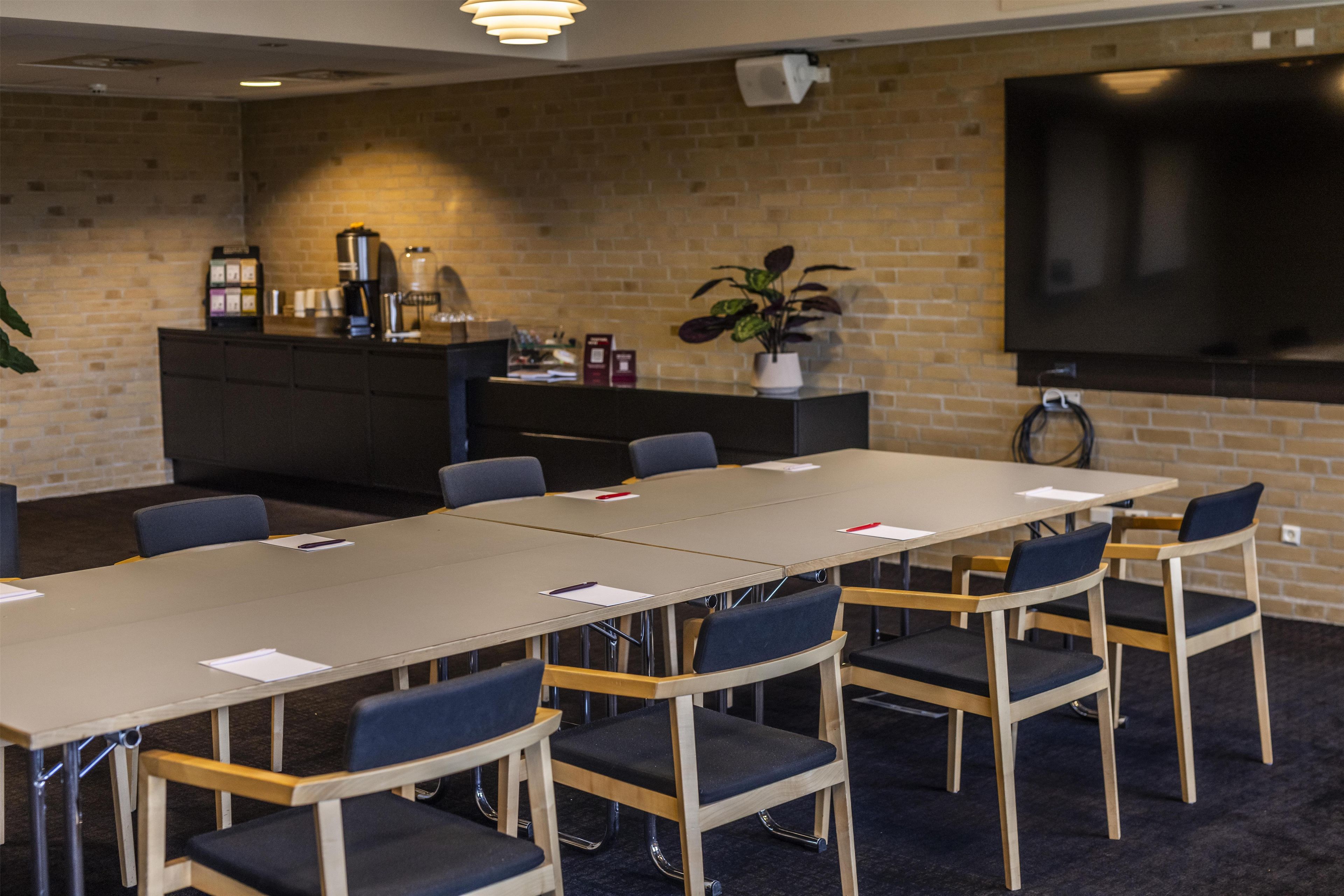 A conference room with a long table, chairs, a coffee machine, a plant, and a TV on the wall.