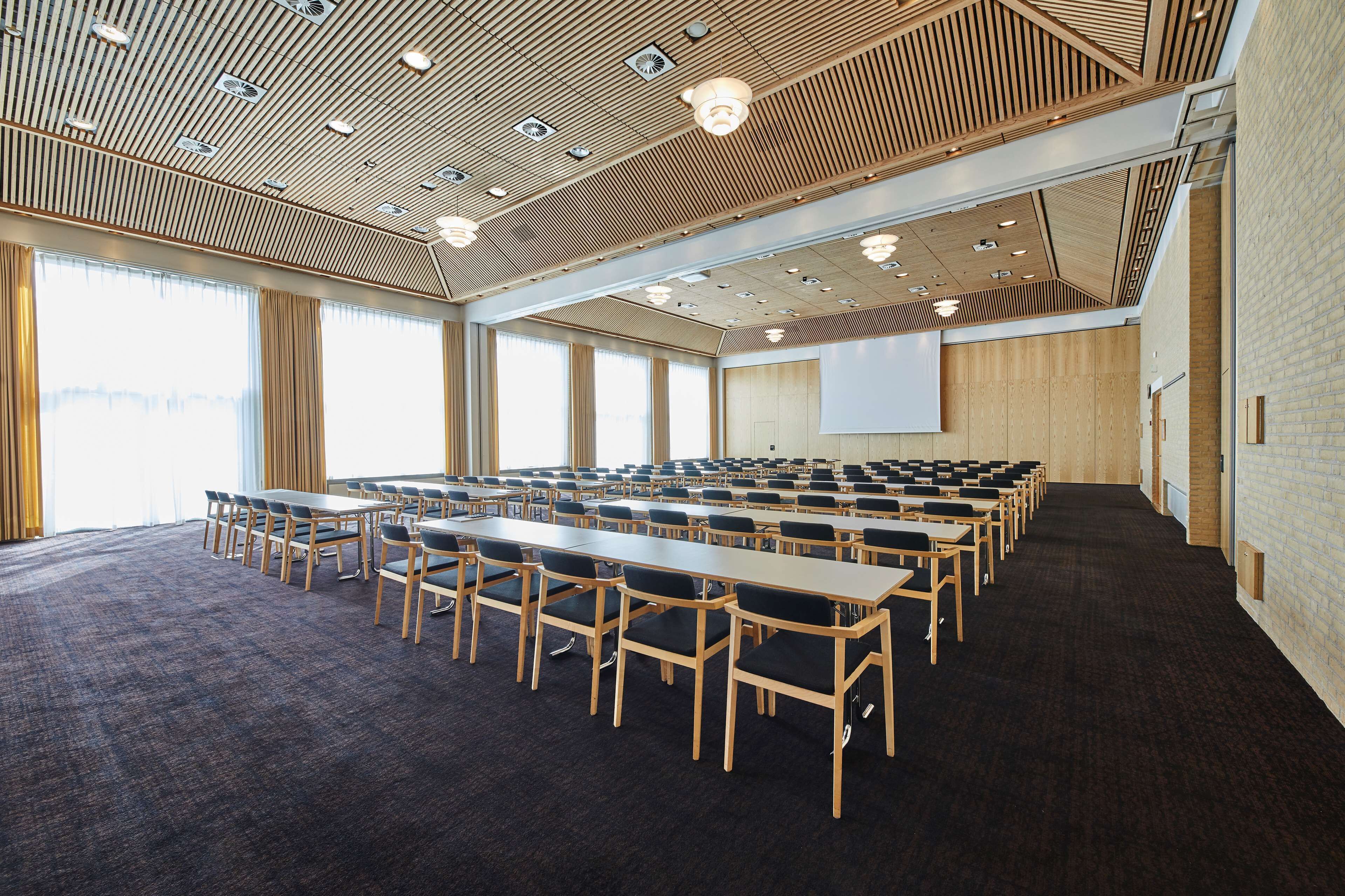 Un salón de conferencias con largas mesas y sillas, una pantalla de proyector y amplias ventanas.