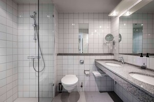 Un baño moderno con paredes de azulejos blancos, cabina de ducha de vidrio, inodoro y tocador con dos lavamanos y un espejo grande.