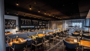 Un restaurante moderno con cabinas y mesas, una zona de bar con botellas de vino y vistas a la ciudad a través de amplias ventanas.