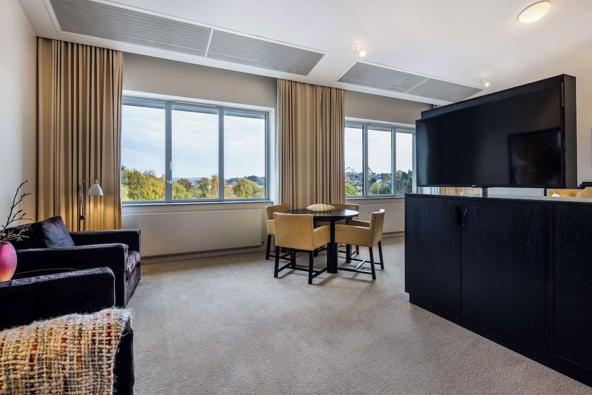 Une chambre d’hôtel moderne dotée d’un coin repas, de grandes fenêtres, d’un téléviseur à écran plat et d’un meuble noir.