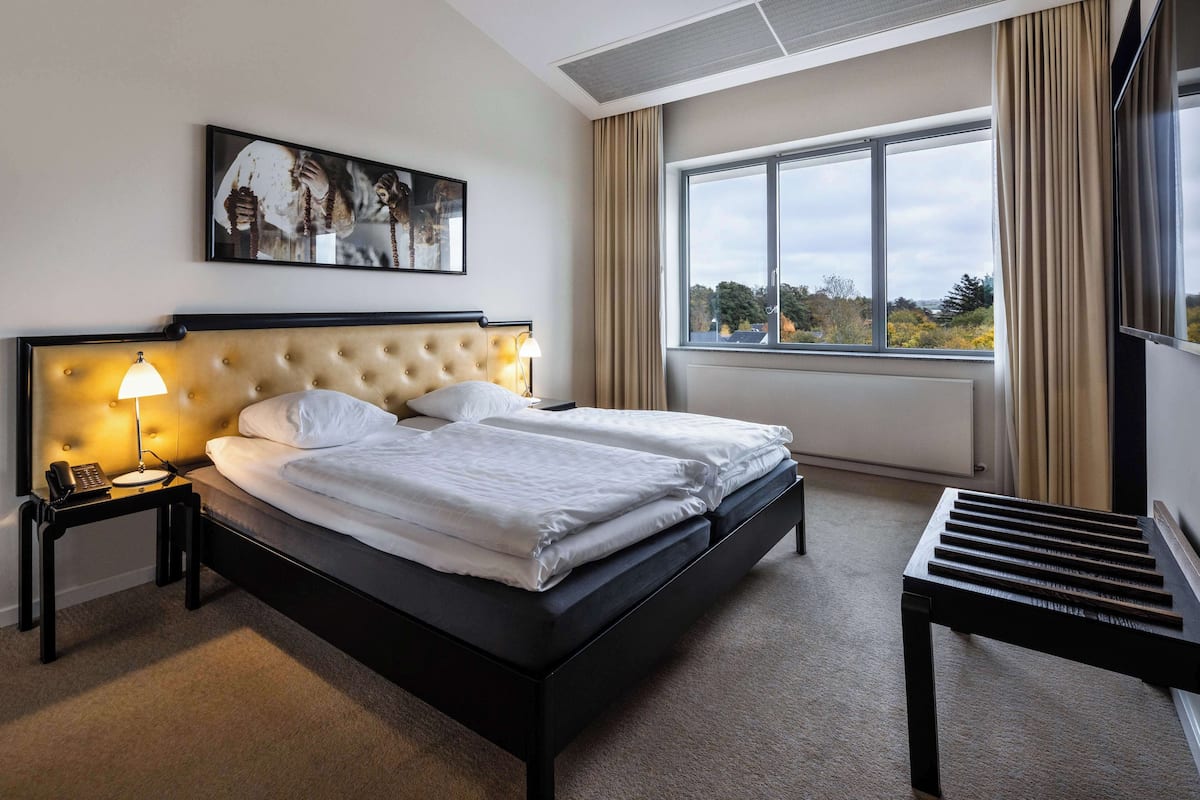 Une chambre d’hôtel avec un grand lit, une table de chevet avec une lampe, une petite table avec un clavier et une fenêtre avec des rideaux.