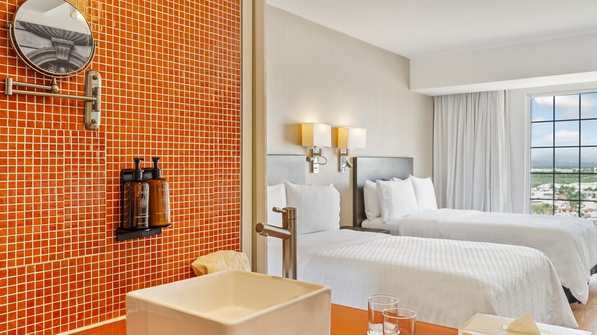 Habitación de hotel con dos camas, un baño con espejo y vistas a la ciudad.