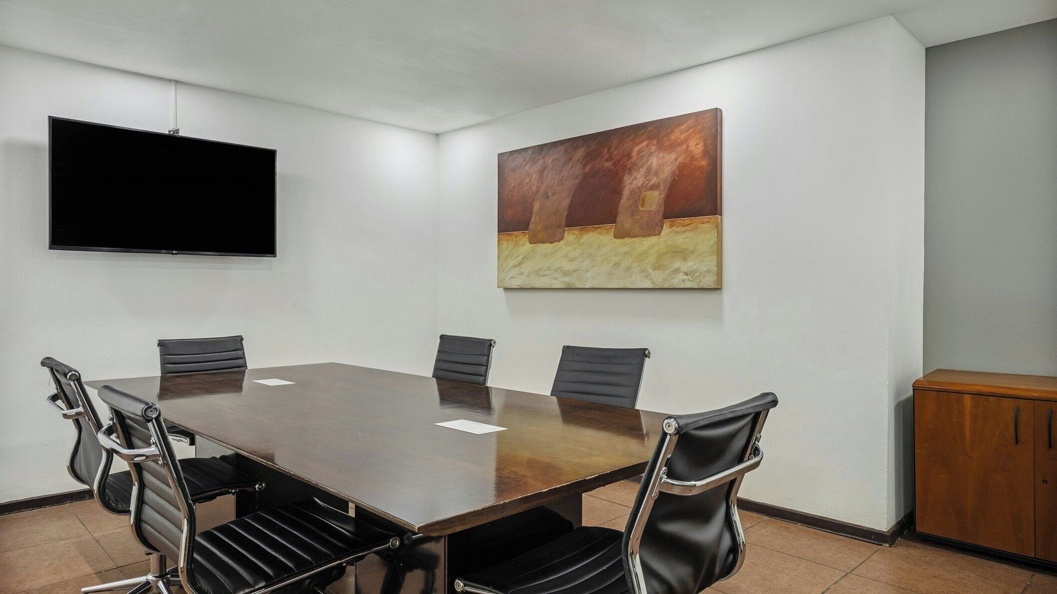 Ein Konferenzraum mit einem langen Tisch, sechs Stühlen, einem Flachbildfernseher und einem großen abstrakten Gemälde an der Wand.