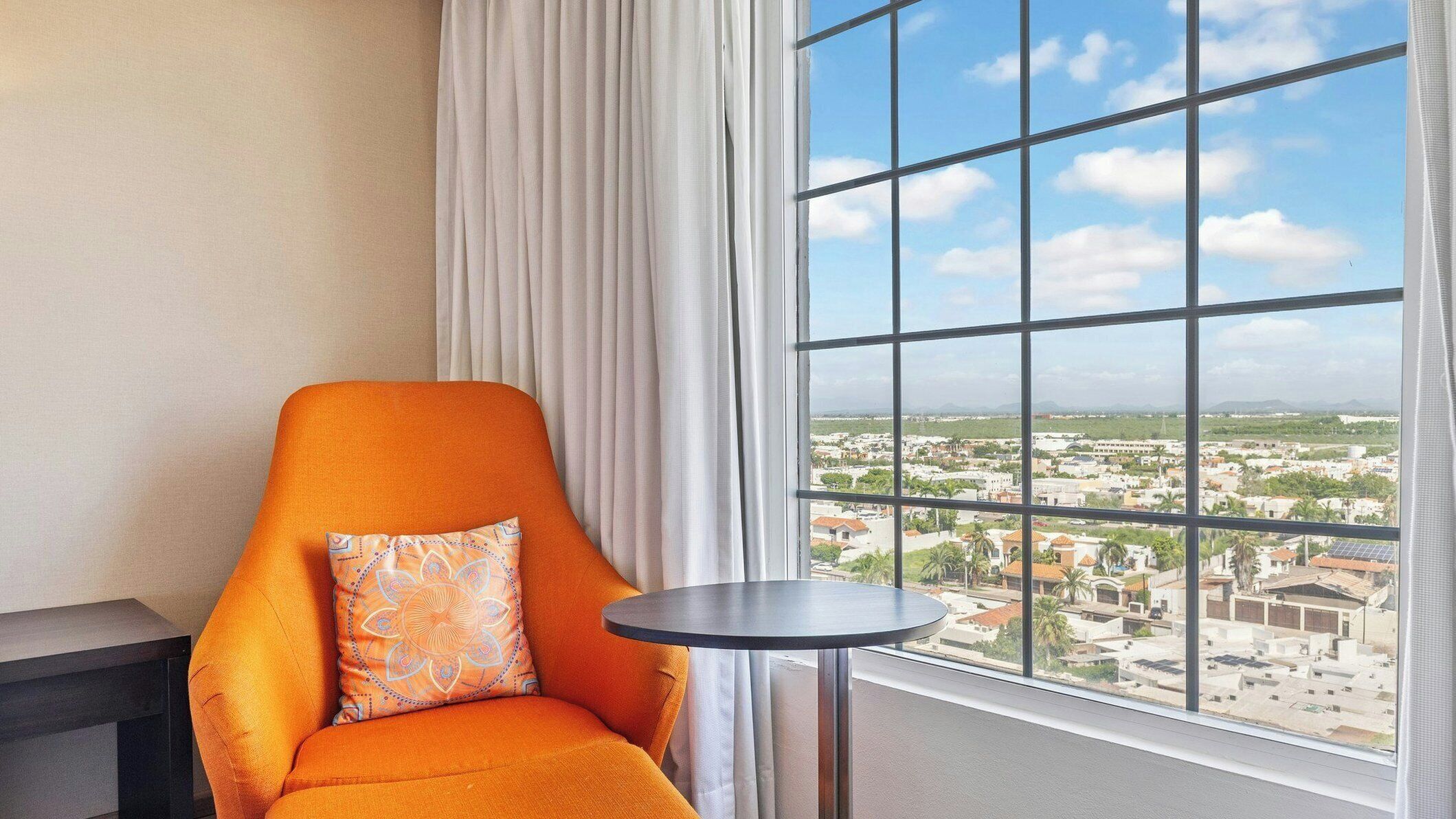 Una habitación con vista a un paisaje urbano a través de un gran ventanal, una silla con cojín y una pequeña mesa redonda.