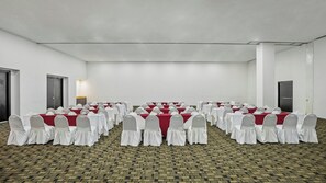 Un salón de conferencias con filas de mesas y sillas, piso alfombrado y paredes blancas.