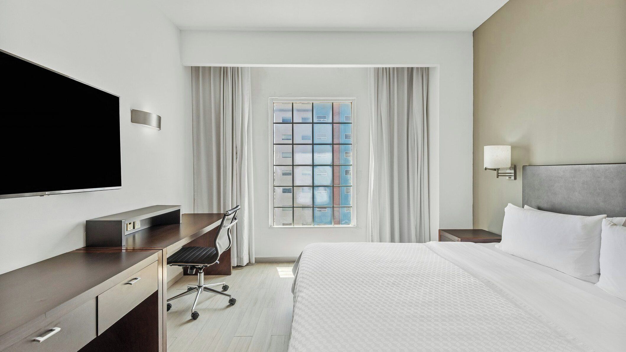 Habitación de hotel con cama, escritorio, televisión, ventana con cortinas y mesita de noche.