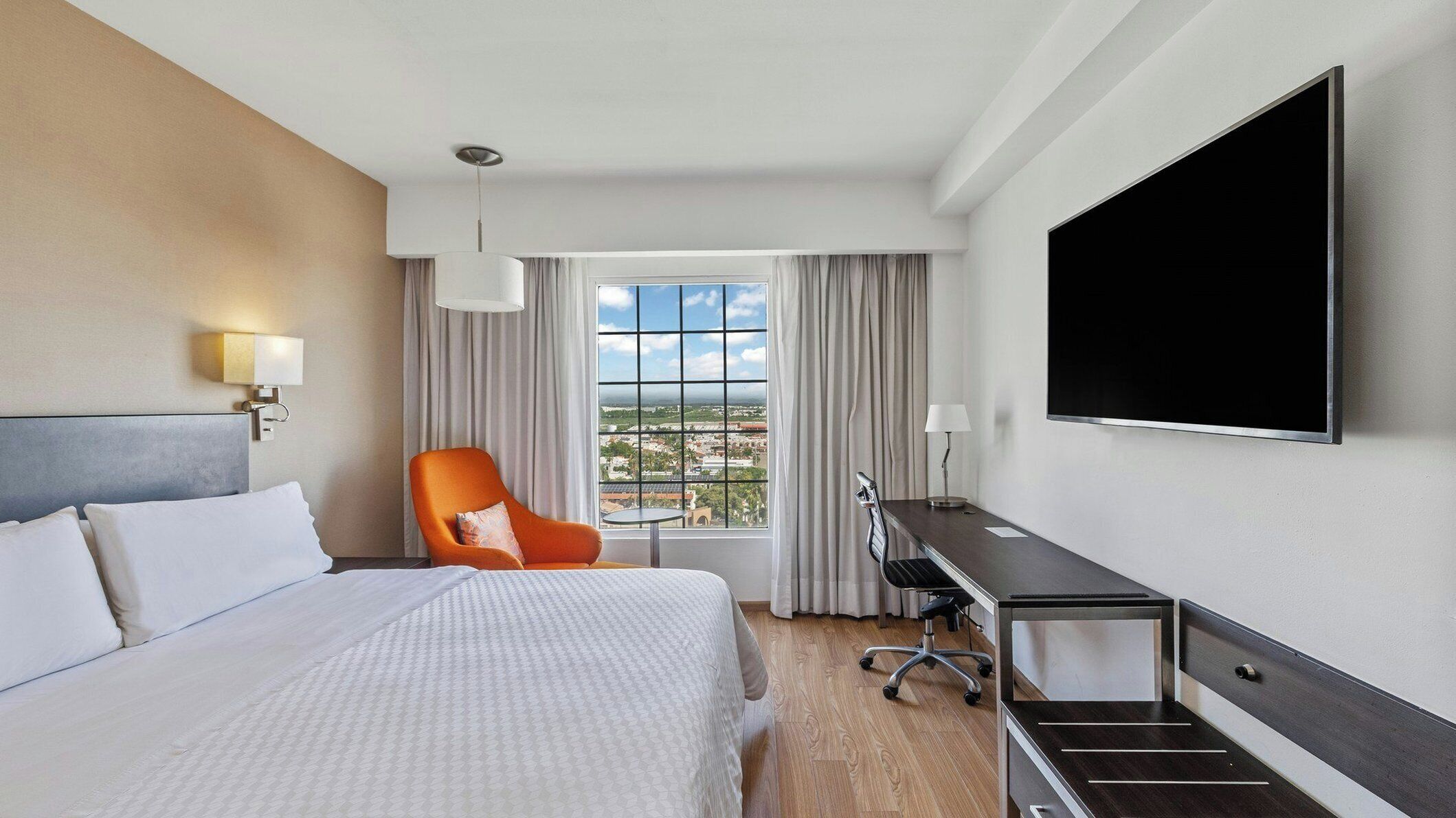 Habitación de hotel con cama, escritorio, silla, televisor y ventana con vistas a la ciudad.