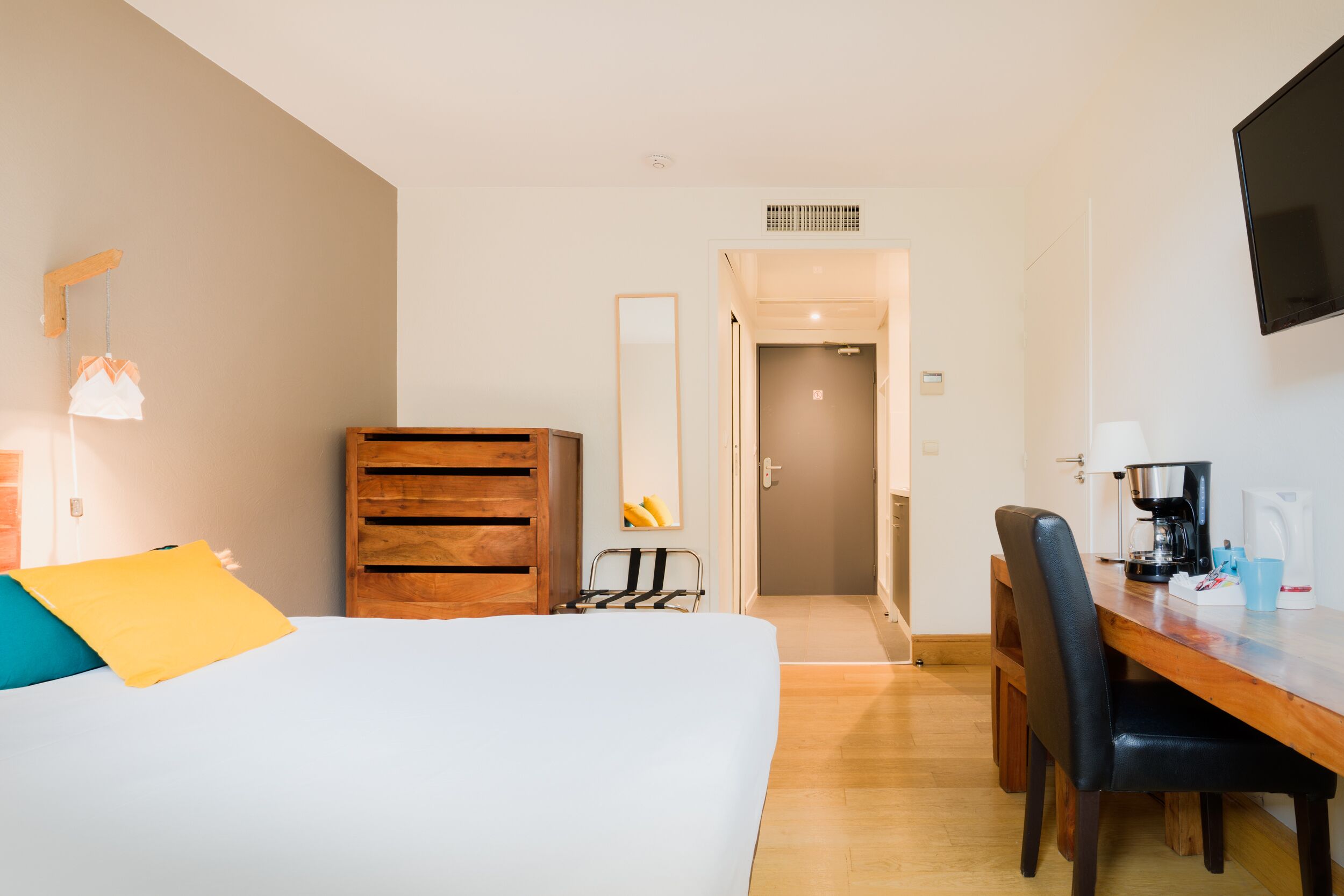 Una habitación de hotel con una cama, un aparador de madera, un escritorio con una silla, una cafetera y un televisor montado en la pared.