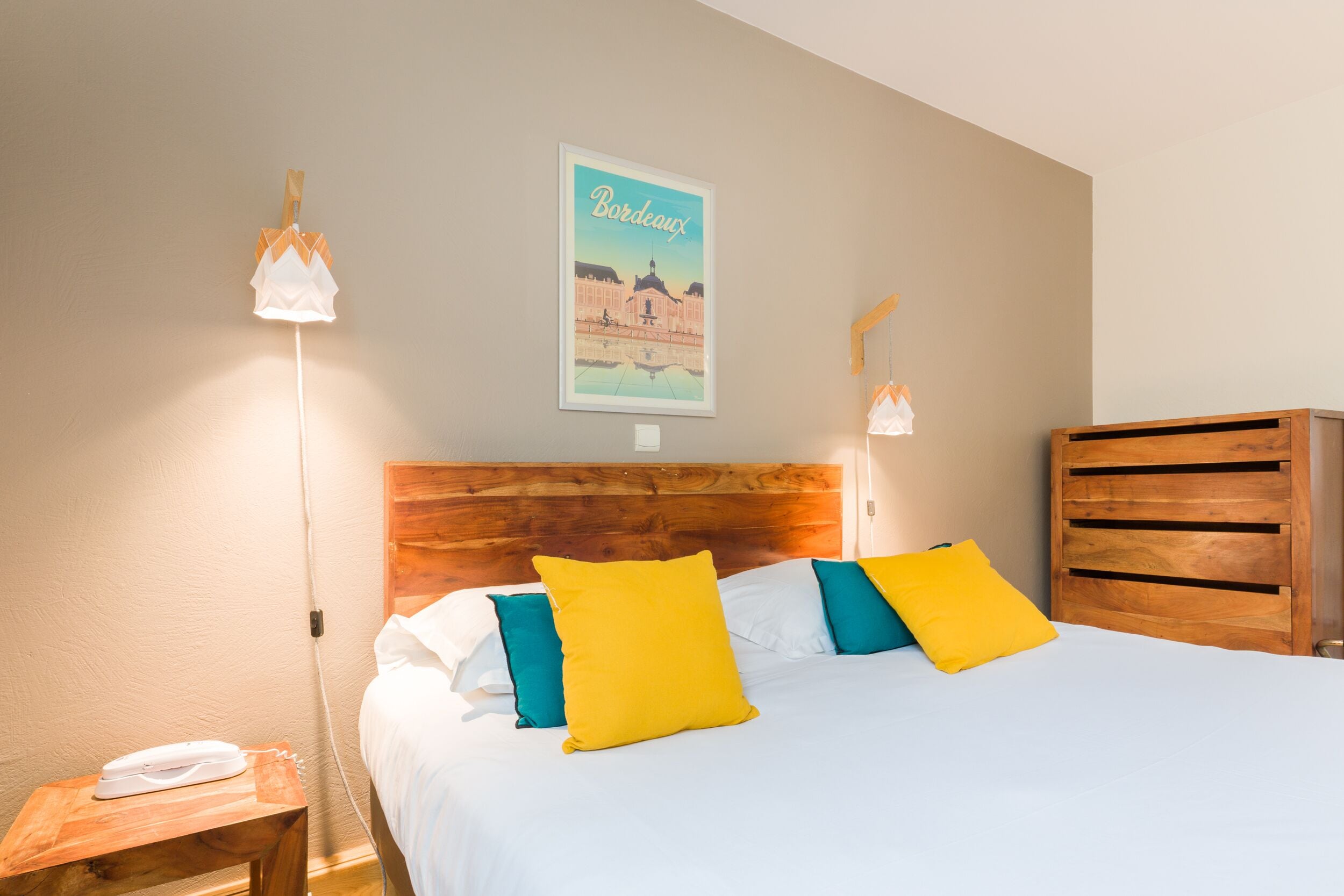 Una cama bien hecha con almohadas blancas y amarillas, un cabecero de madera y un cartel de Bordeaux en la pared.