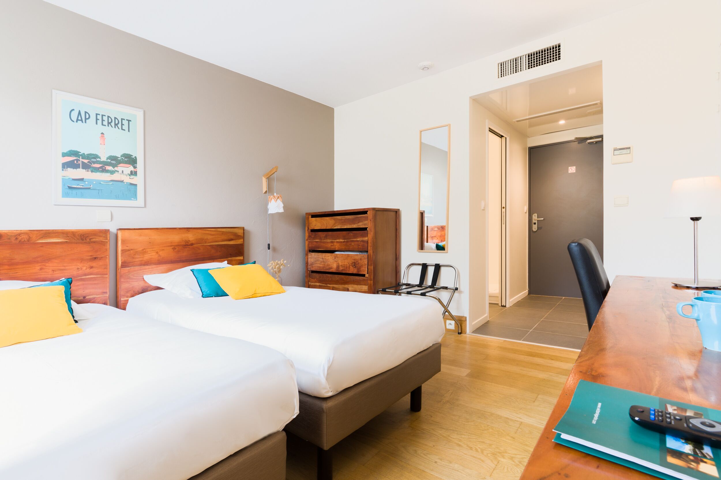 Quarto de hotel com duas camas, uma cômoda de madeira, uma mesa de trabalho com controle remoto e um quadro na parede.