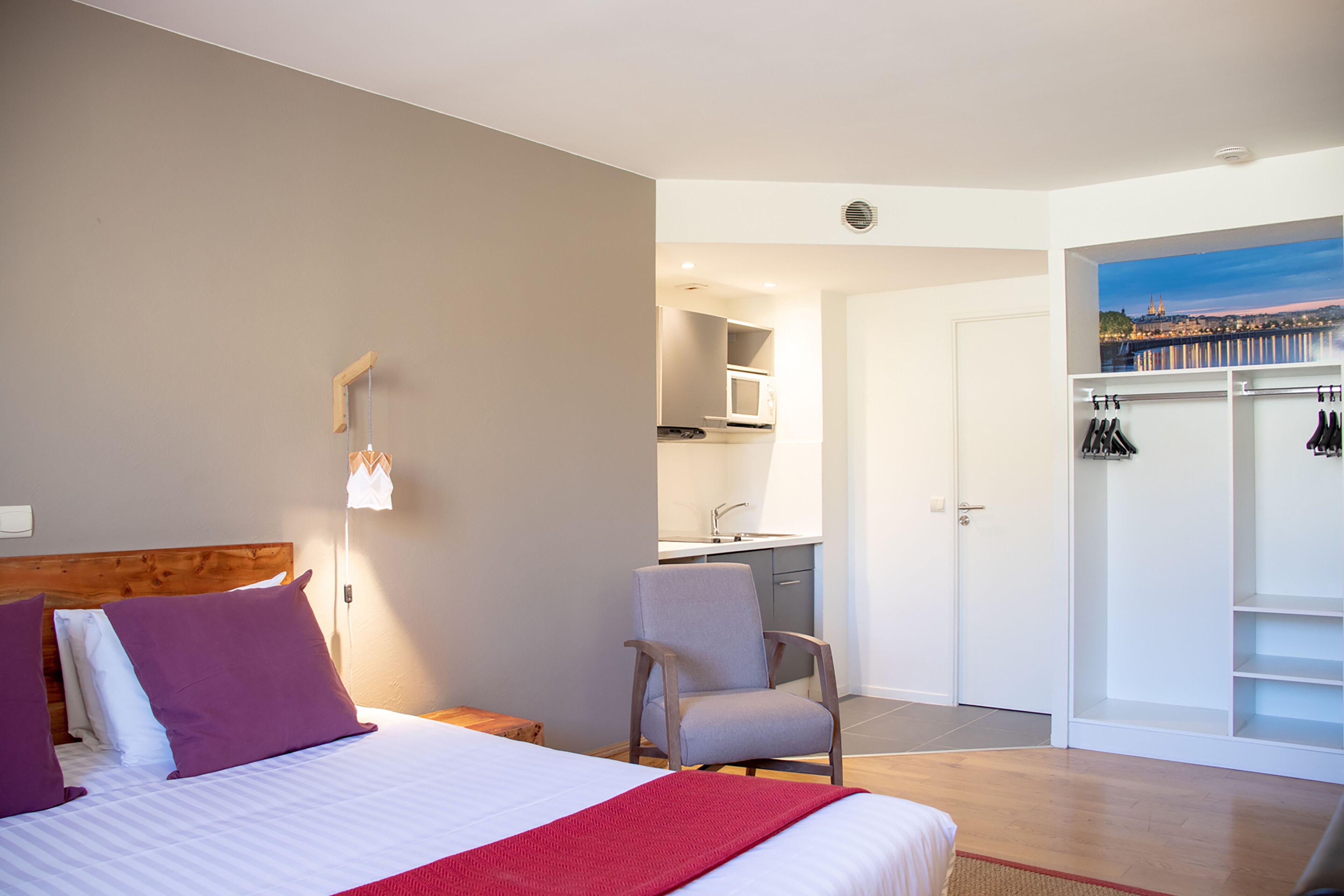 A hotel room with a bed, a chair, a wardrobe, and a view of a cityscape.