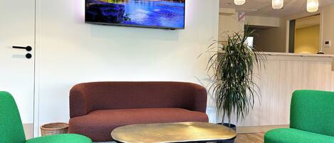Una moderna zona de recepción con un sofá, sillas verdes, una mesa redonda y un televisor que muestra un paisaje escénico.