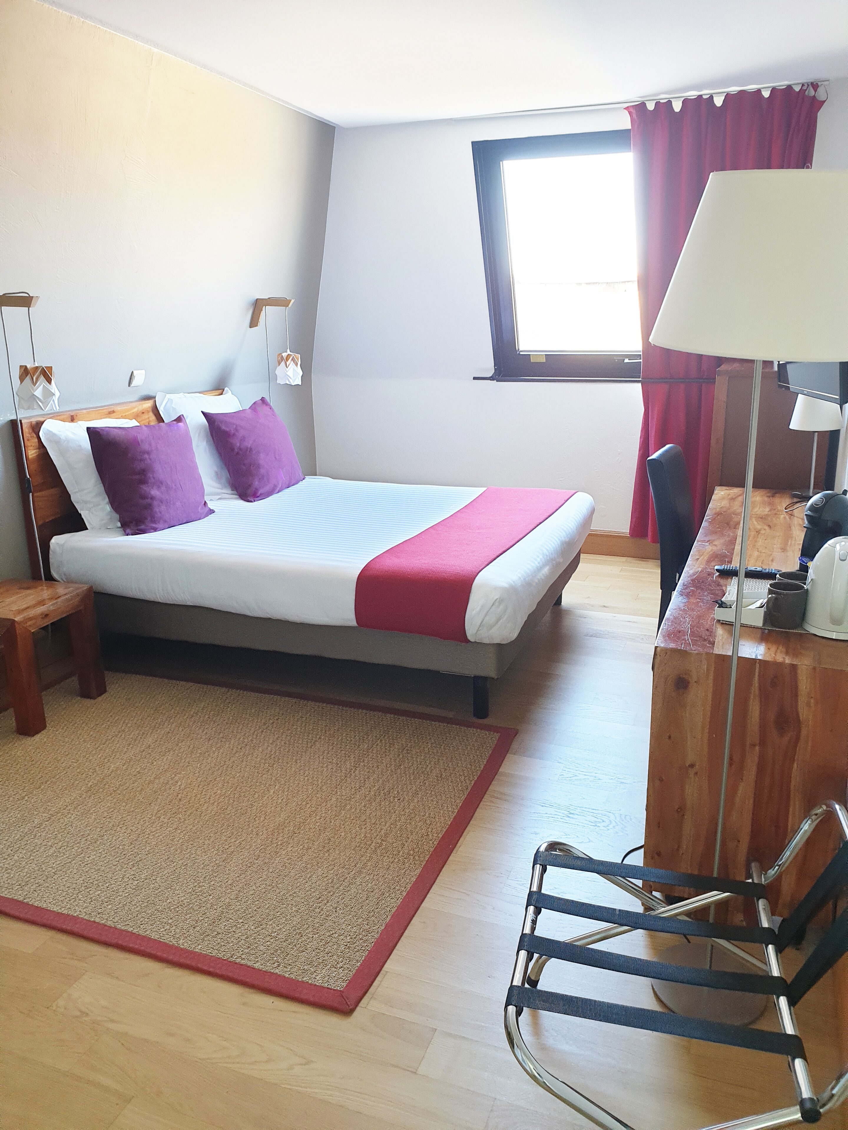 Une chambre d’hôtel avec un lit, un bureau, une chaise, une fenêtre avec des rideaux et une lampe.