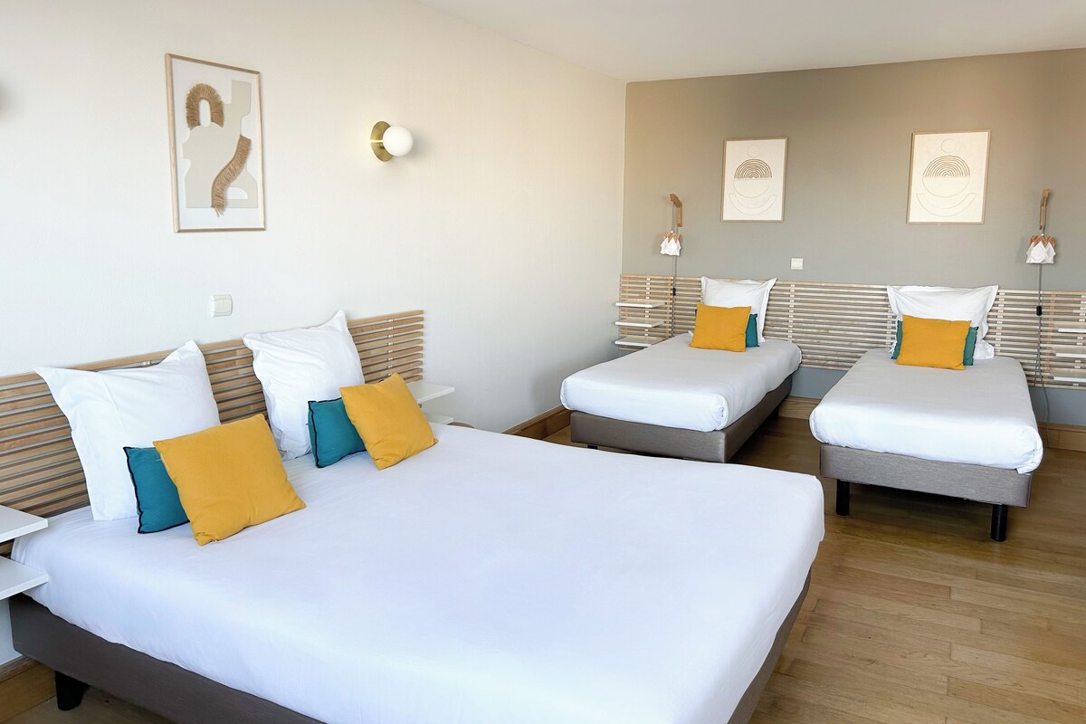 Habitación de hotel con dos camas, cada una con ropa blanca y varias almohadas. Hay lámparas de pared y cuadros enmarcados sobre las camas.
