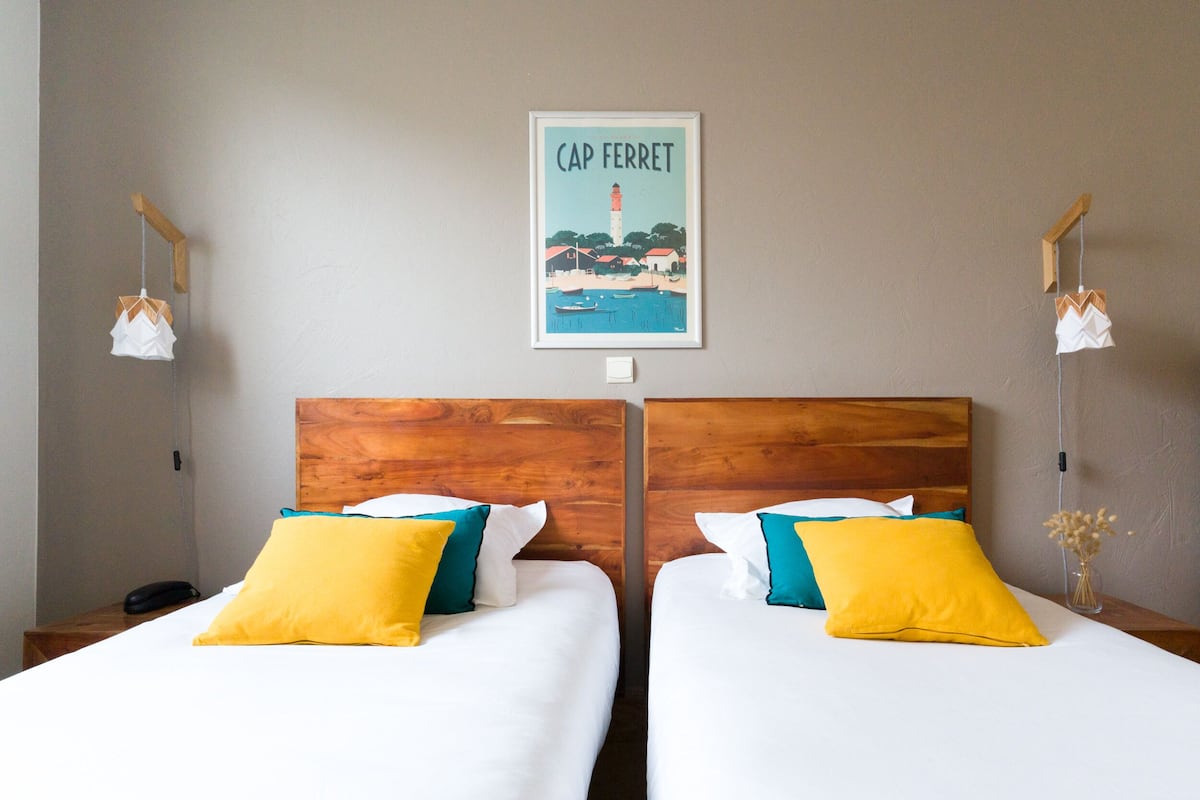 Hotelkamer met twee bedden, elk voorzien van witte beddengoed en gele kussens, een houten hoofdbord, een lijst met een afbeelding van Cap Ferret, en bedlampjes.