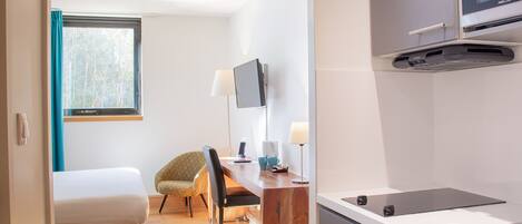 A modern hotel room with a bed, a desk with a chair, a TV, and a kitchenette.