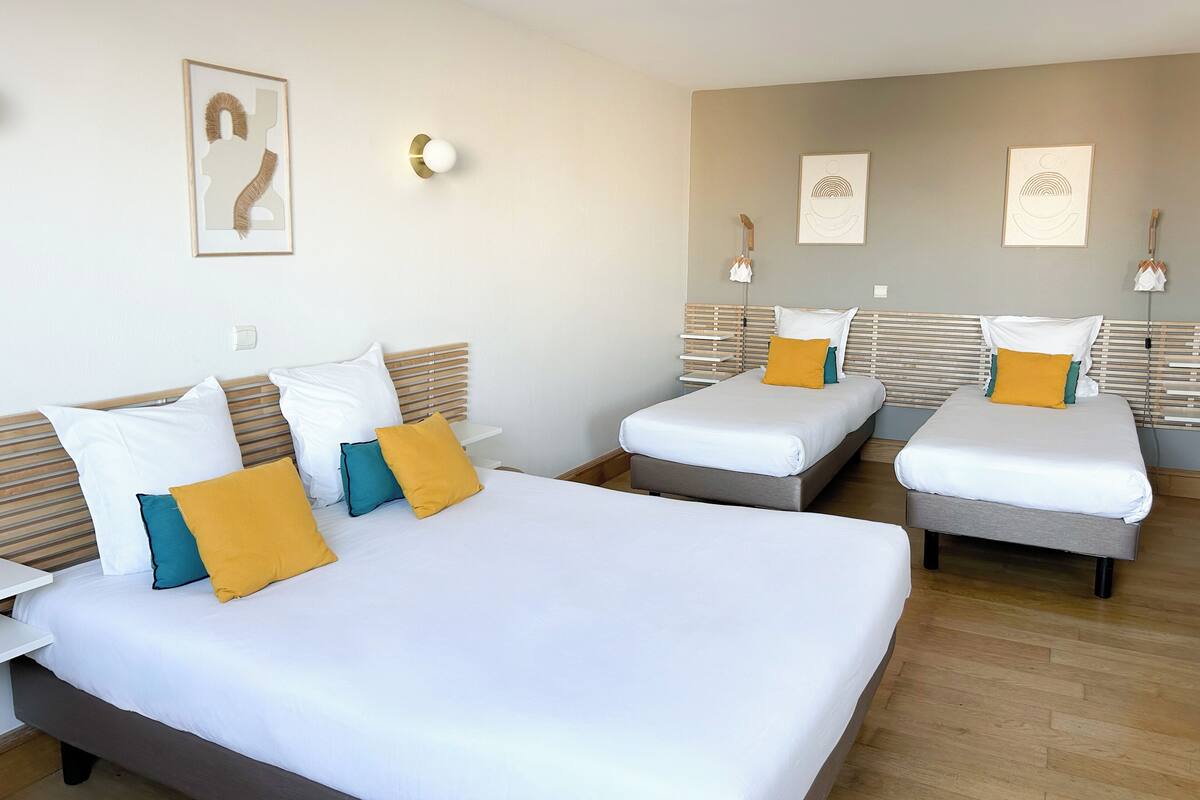 Een hotelkamer met twee eenpersoonsbedden en een groter bed, elk voorzien van witte lakens en kleurrijke kussens. Er hangen wandlampen en kunstwerken boven de bedden.