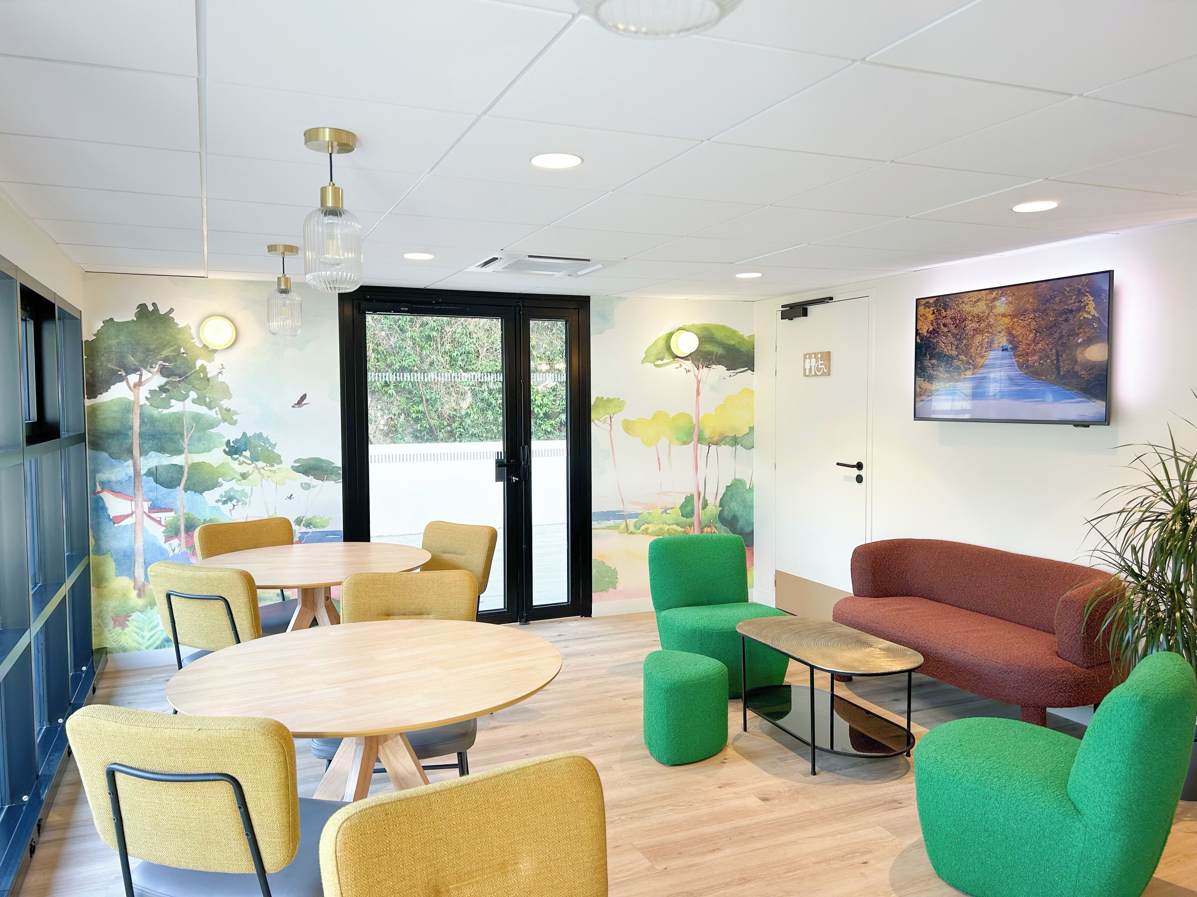 A modern meeting room with round tables, green and yellow chairs, a red sofa, and a large window with a view of trees and a road.