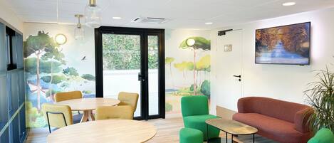 A modern meeting room with round tables, green and yellow chairs, a red sofa, and a large window with a view of trees and a road.