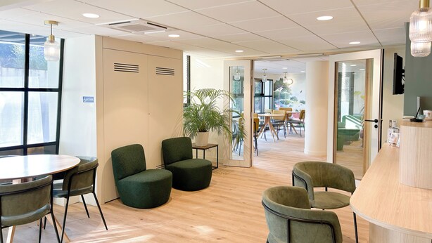 Un espacio de oficina moderno con una mesa redonda, sillas tapizadas en verde, un mostrador de recepción y un ventanal grande.