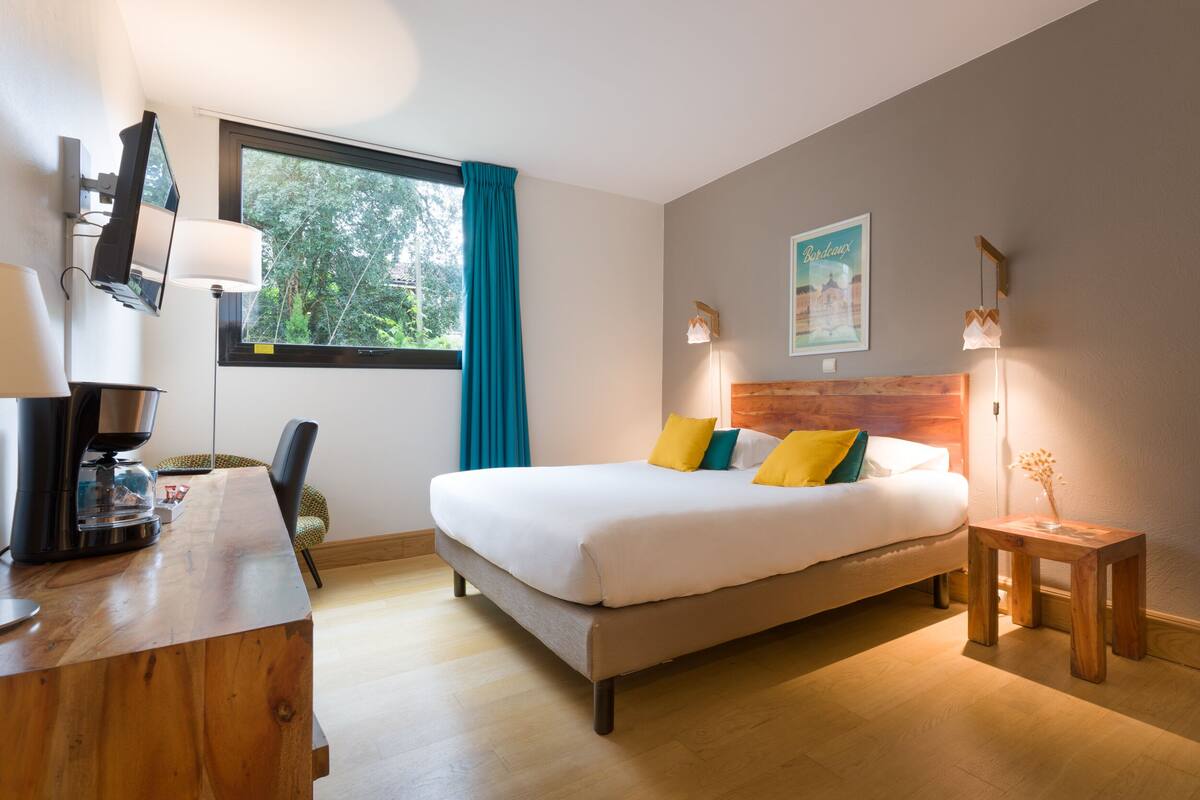 Een moderne hotelkamer met een groot bed, een bureau met koffiezetapparaat, een stoel en een raam met gordijnen.
