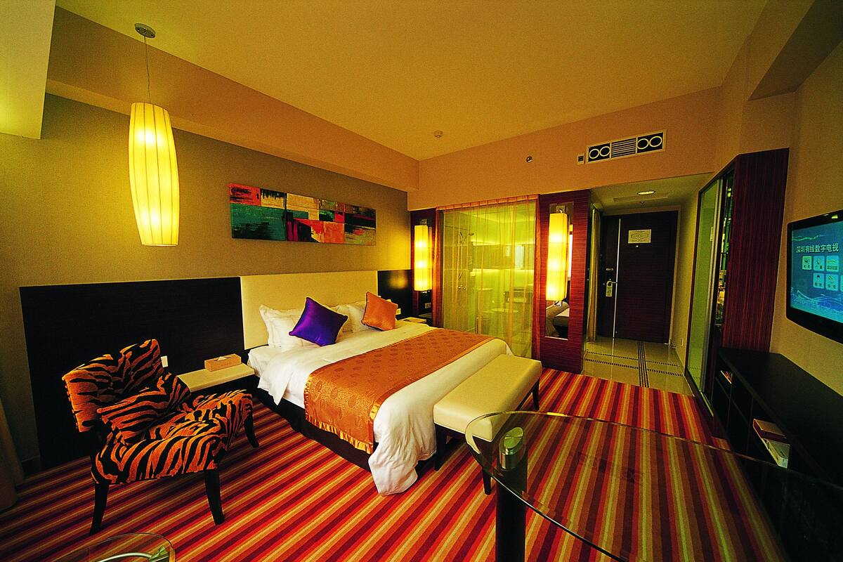 A hotel room with a bed, a chair, a TV, and a glass door leading to a bathroom.
