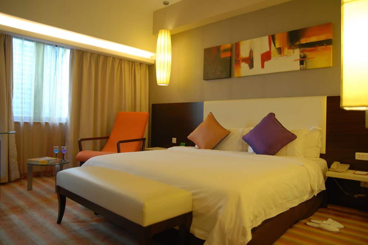 A hotel room with a large bed, a chair, a small table with glasses, and a painting on the wall.
