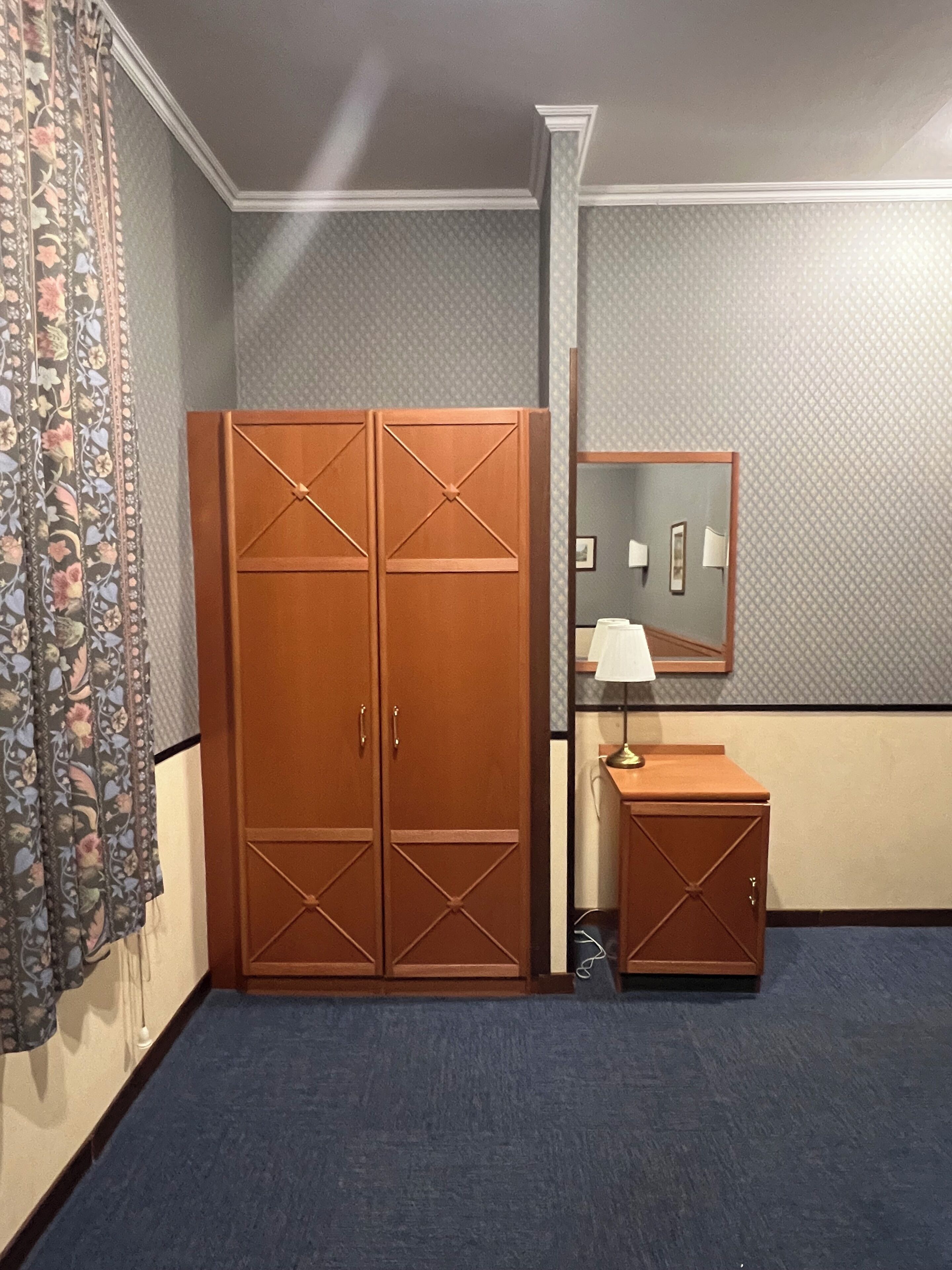 Une chambre avec une grande armoire, une petite table de chevet, un miroir et un rideau à motifs.