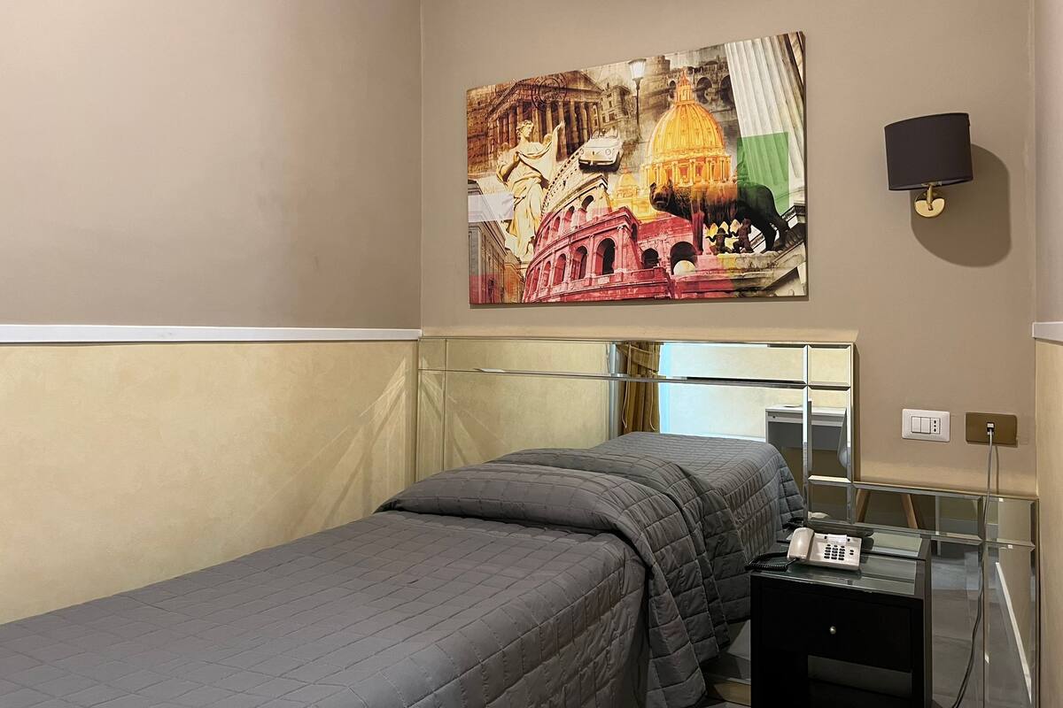 A hotel room with a bed, a painting of a historical building, a bedside table with a lamp, and a mirror.