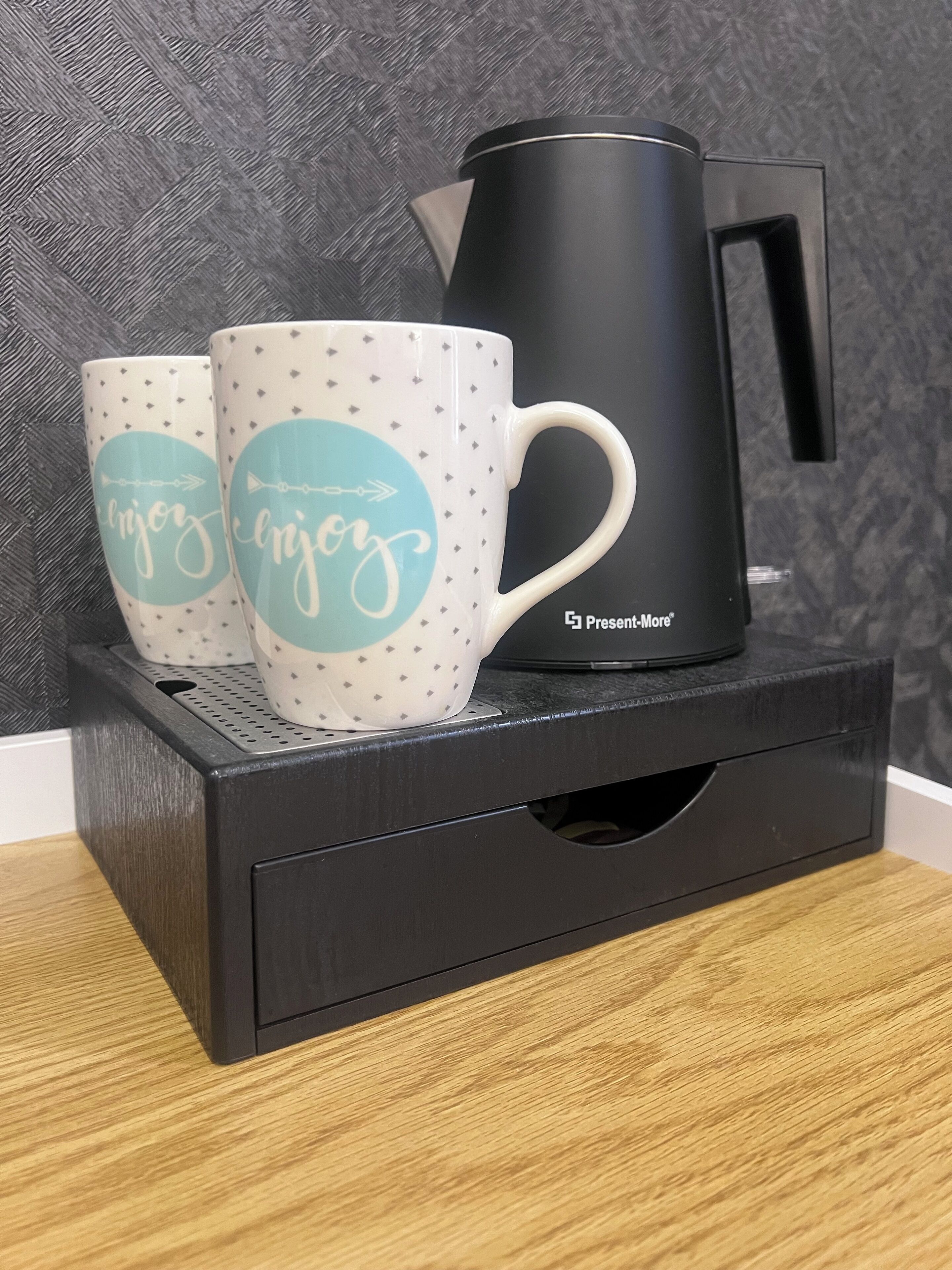 Two white mugs with teal accents and the word "enjoy" on a black coffee maker on a black tray.