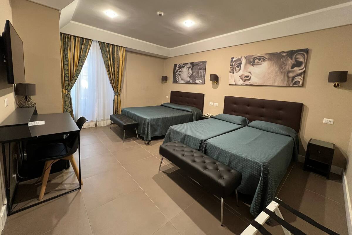 A hotel room with two beds, a desk, a bench, and artwork on the walls.