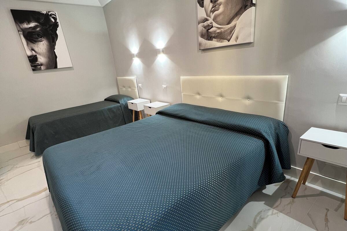 A bedroom with two beds, a nightstand, a desk, and a large wall-mounted artwork.