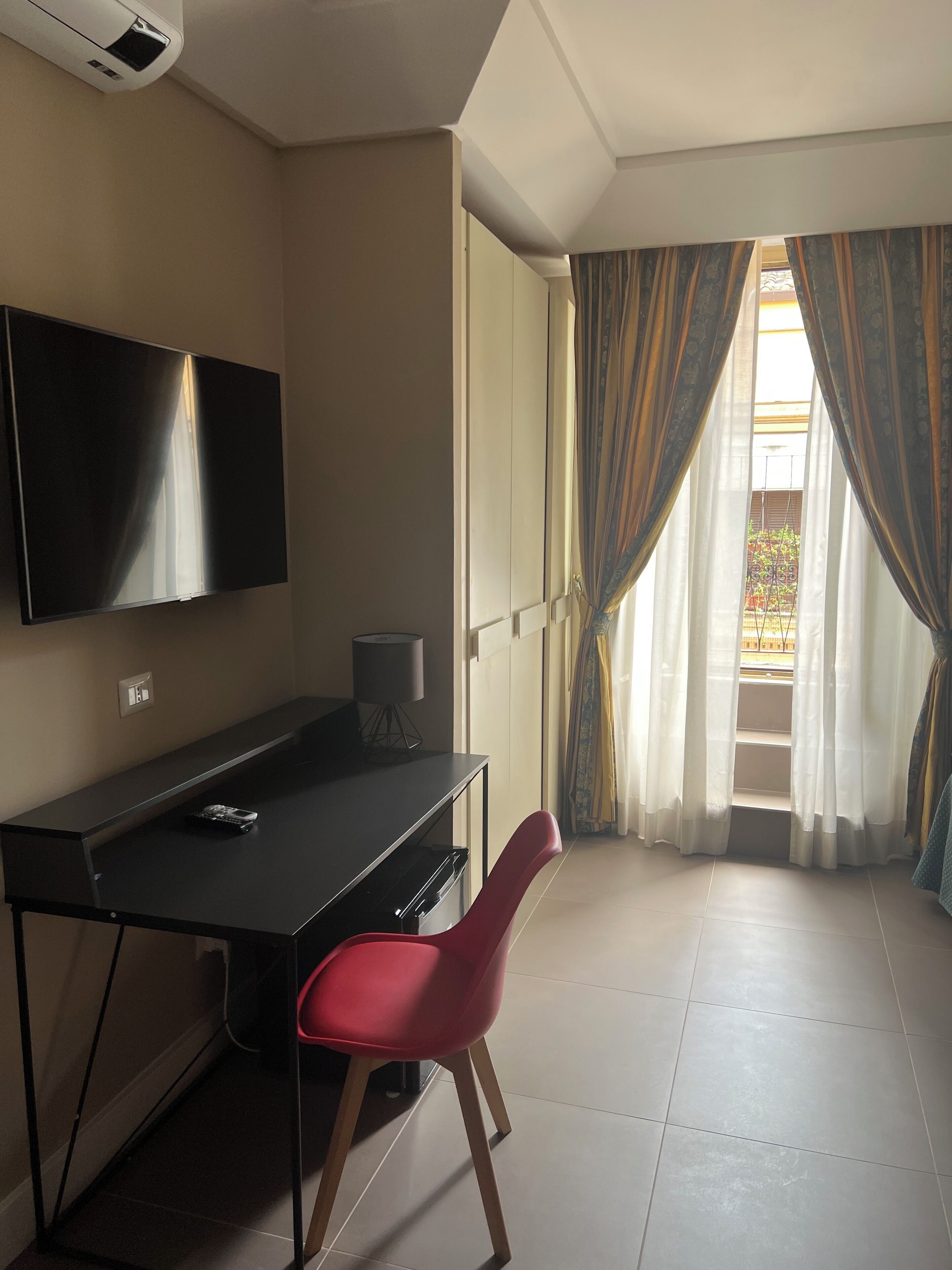 A room with a flat-screen TV mounted on the wall, a desk with a chair, and a window with curtains.