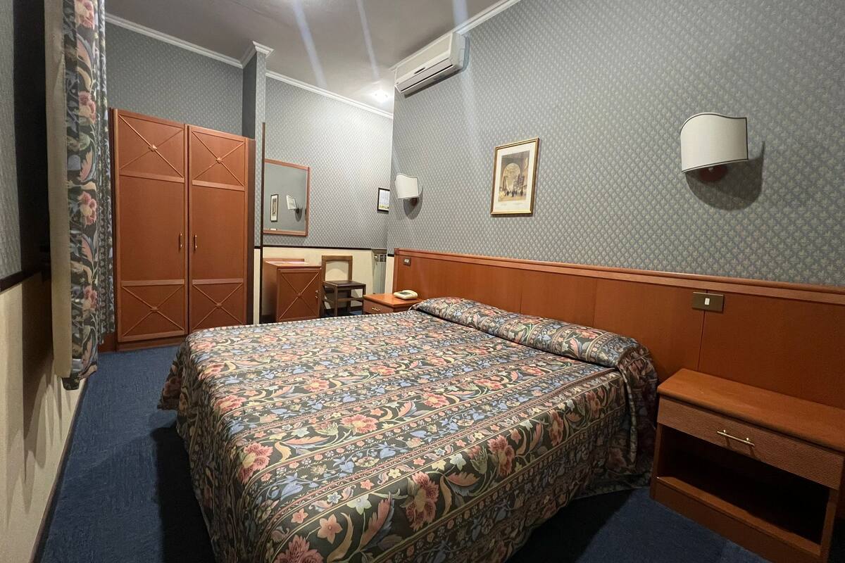 A hotel room with a large bed, a wooden wardrobe, a desk with a telephone, and a mirror.