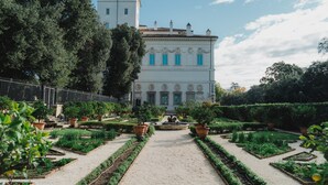 A garden pathway leading to a white building with blue shutters, surrounded by neatly arranged plants and trees. - Hotel Garda (Rome)