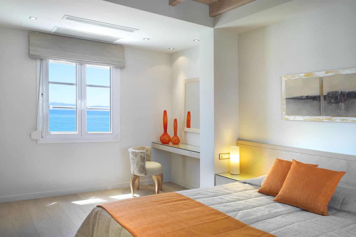 A bedroom with a bed, a nightstand, a chair, a window with a view of the sea, and a painting on the wall.