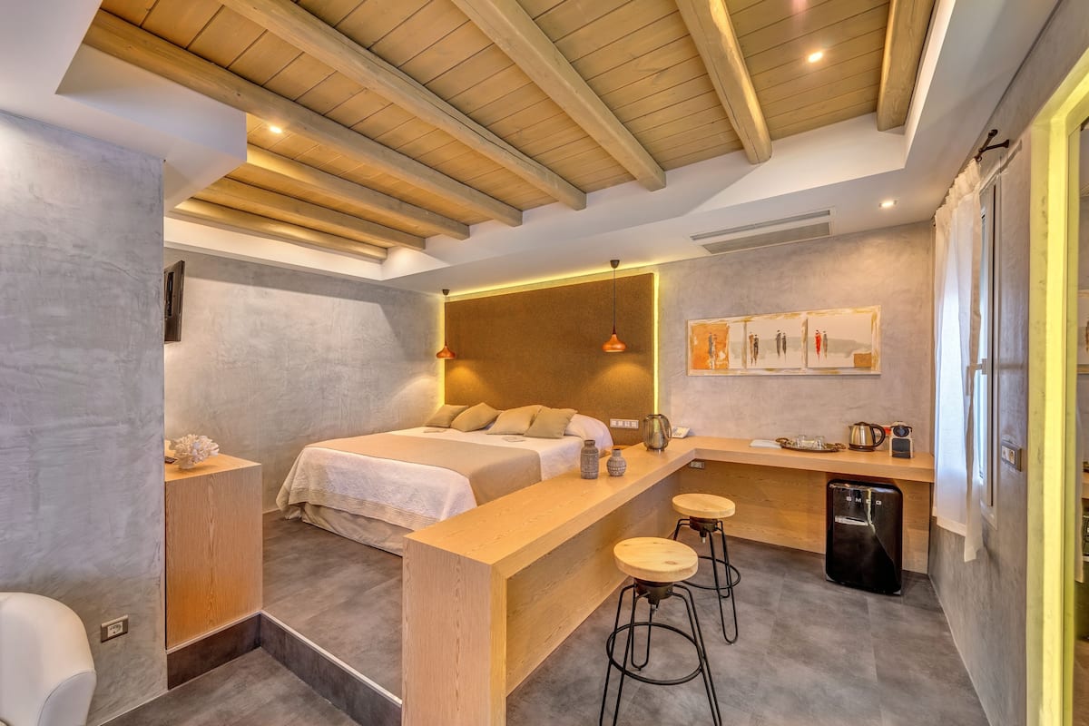 A modern bedroom with a wooden ceiling, a bed with a headboard, a small kitchen area with stools, and a refrigerator.