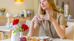 Una persona sentada a una mesa, con una taza, un plato de comida y un jarrón con flores.