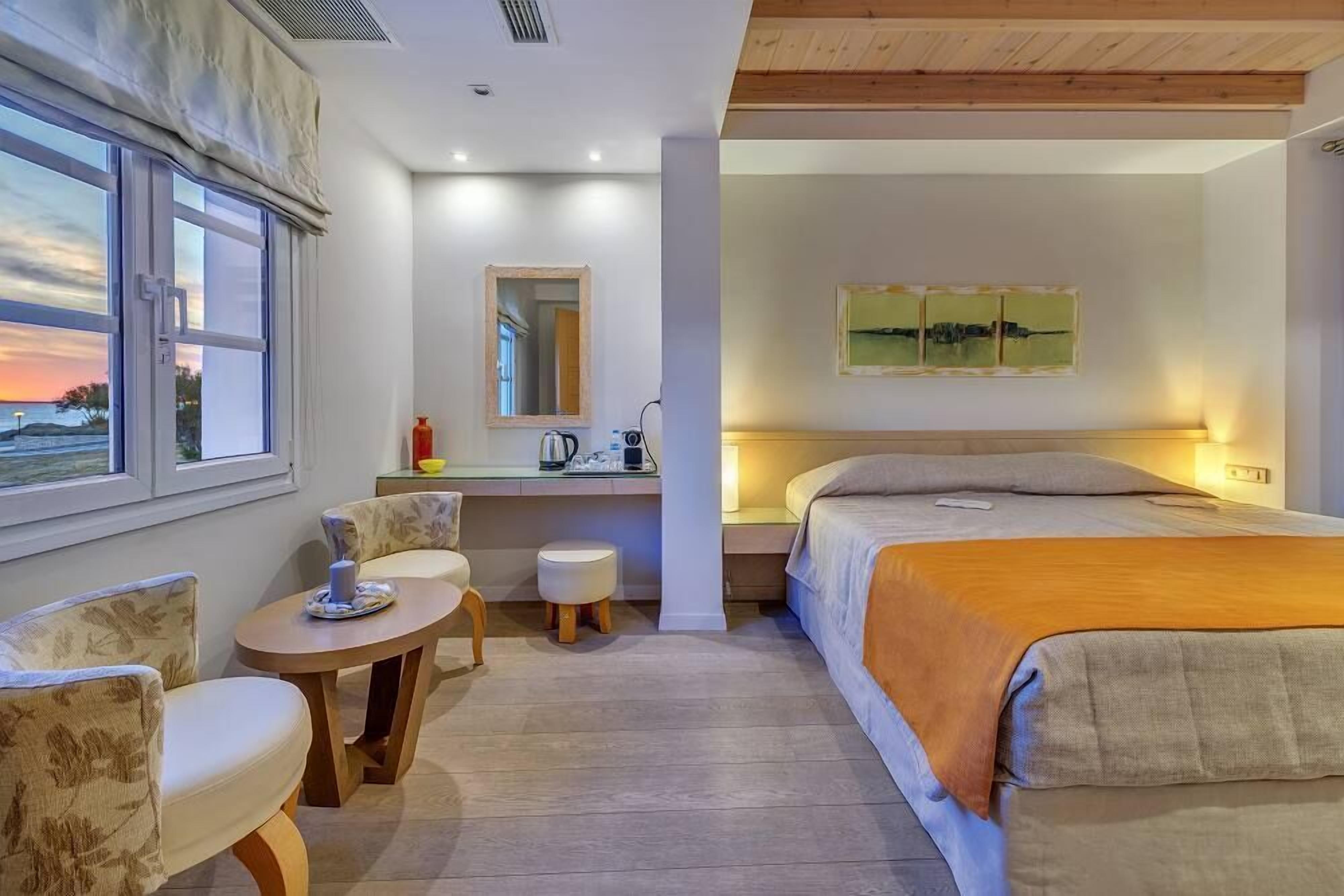 A hotel room with a bed, a desk with a chair, a small table, and a window with a view of the sunset.