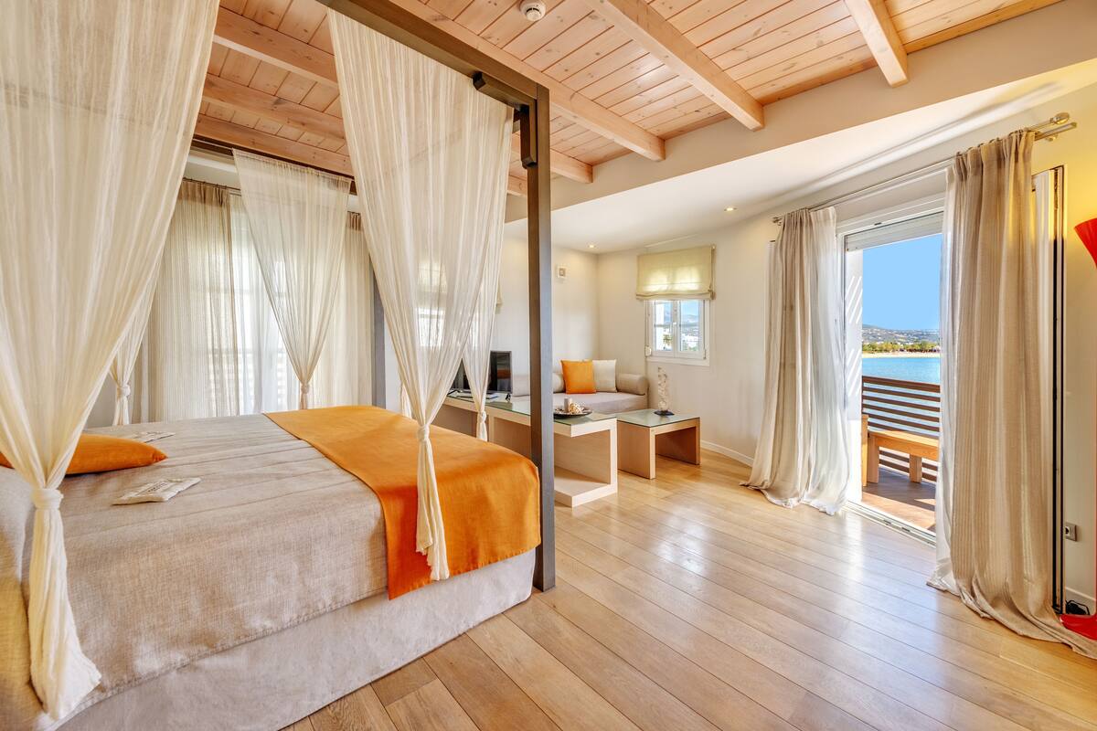 A bedroom with a bed, curtains, a desk, and a view of the sea.